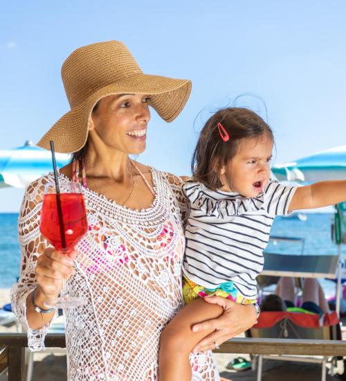 Woman with hat and child on beach, holding a cocktail.